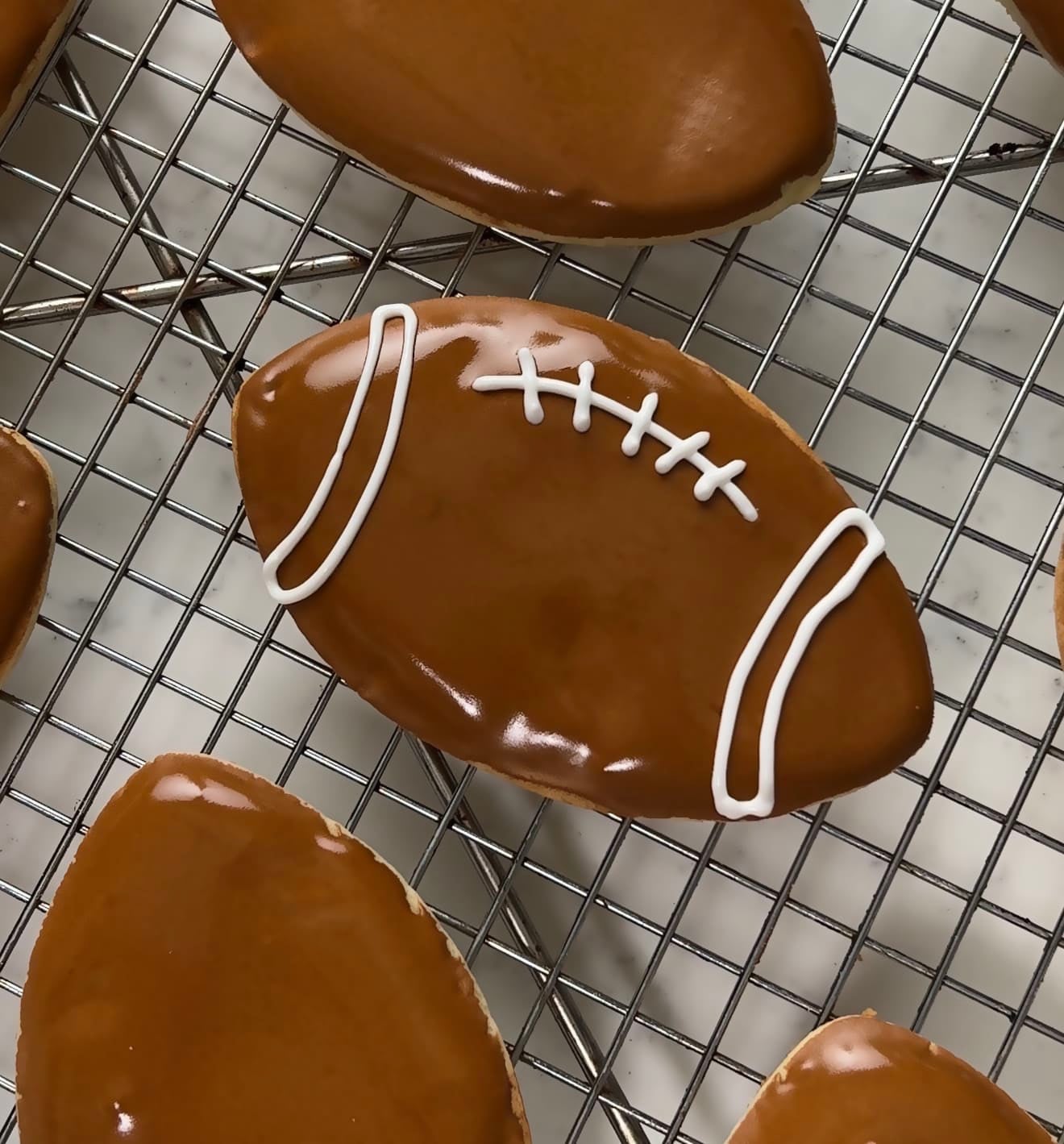 Football sugar cookies dipped in glossy brown royal icing with piped white laces on a cooling rack.
